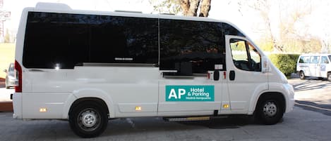 Ein weißer Van mit der Aufschrift "AP Hotel & Parking Madrid Aeropuerto", geparkt auf einer Betonfläche, im Hintergrund Bäume und weitere Fahrzeuge.