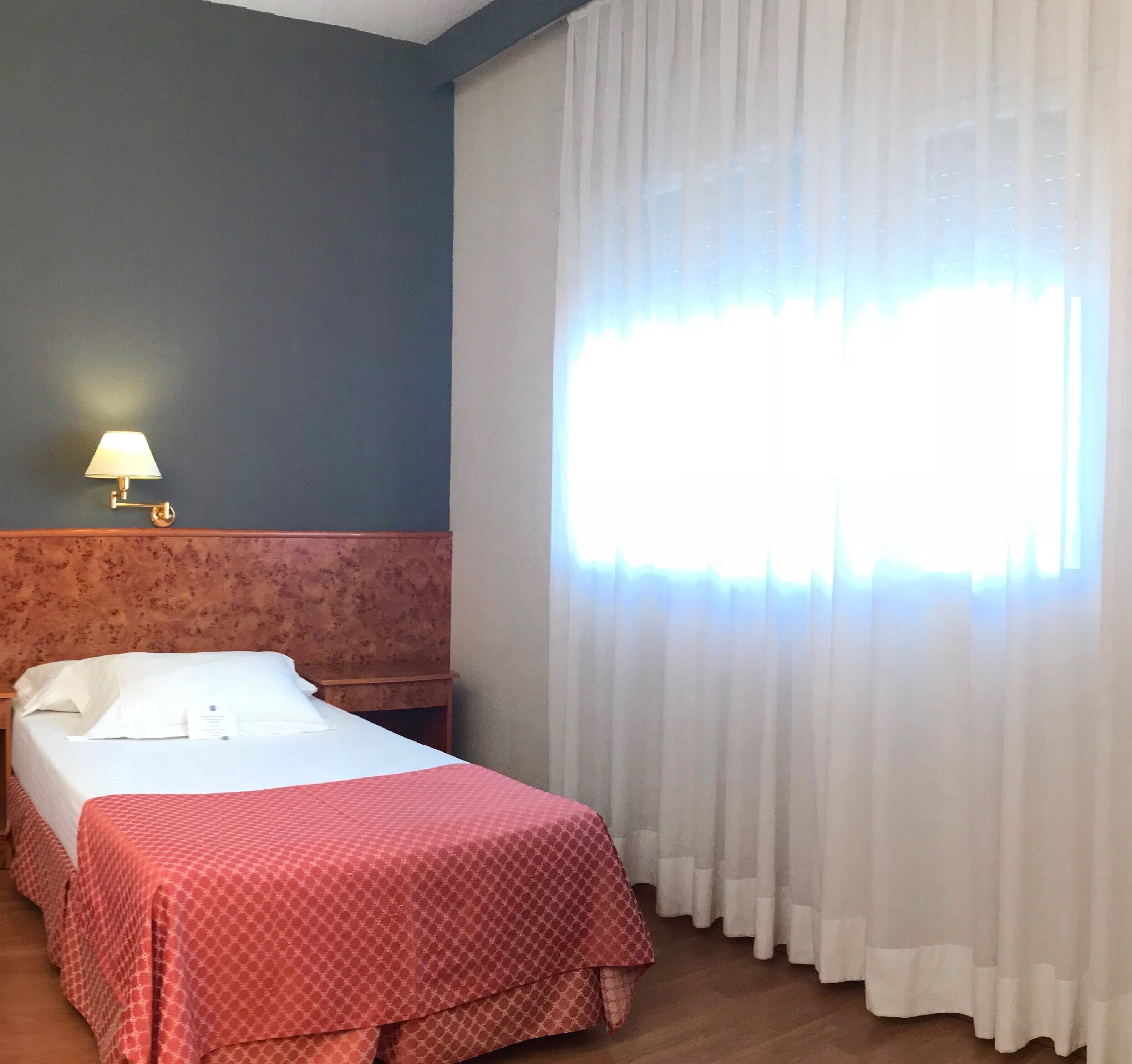 Una habitación de hotel con cama, mesita de noche, lámpara y ventana con cortinas.