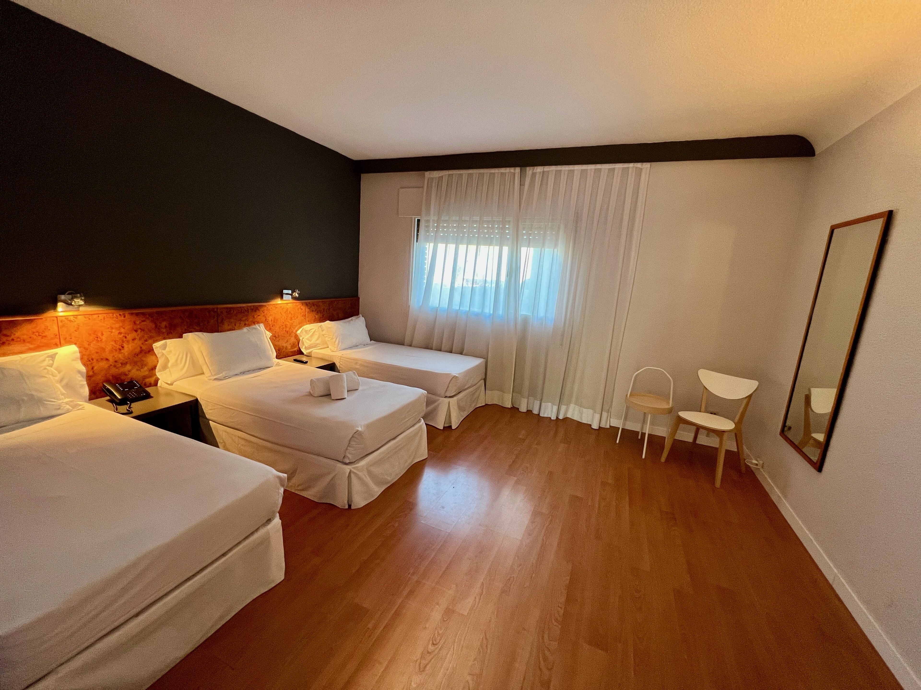 A hotel room with two beds, a small table, a chair, a mirror, and a window with curtains.