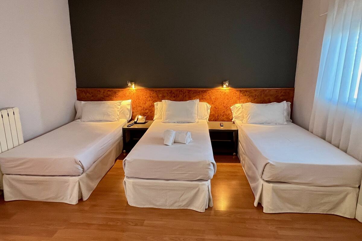 Ein Hotelzimmer mit zwei Einzelbetten, einem Holz-Kopfteil, einem Nachttisch mit Telefon und einem Fenster mit durchsichtigen Vorhängen.