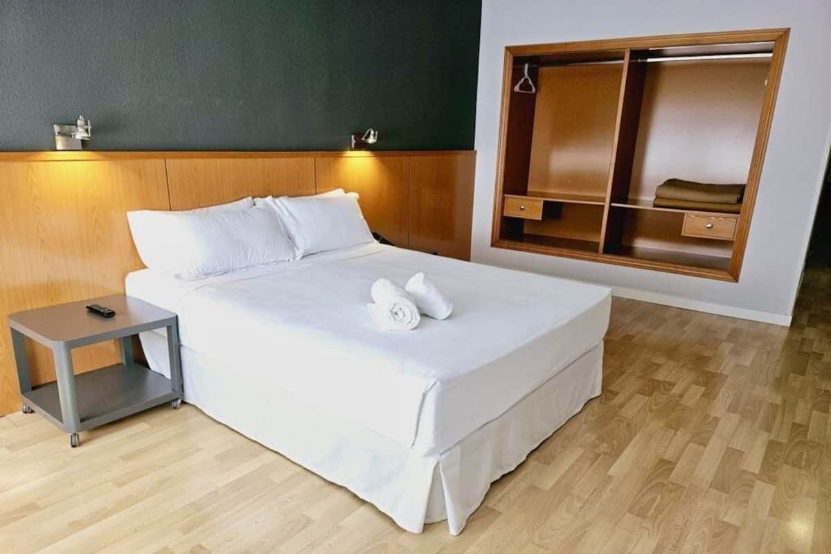 Ein ordentlich bezogenes Bett mit wei ßen Leinen, ein Nachttisch mit Fernbedienung und ein Holzschrank mit Spiegel.