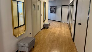 Un couloir doté d’un plancher en bois, de murs blancs et de portes ornées de miroirs. On y trouve un petit banc à carreaux et un tableau encadré accroché au mur.