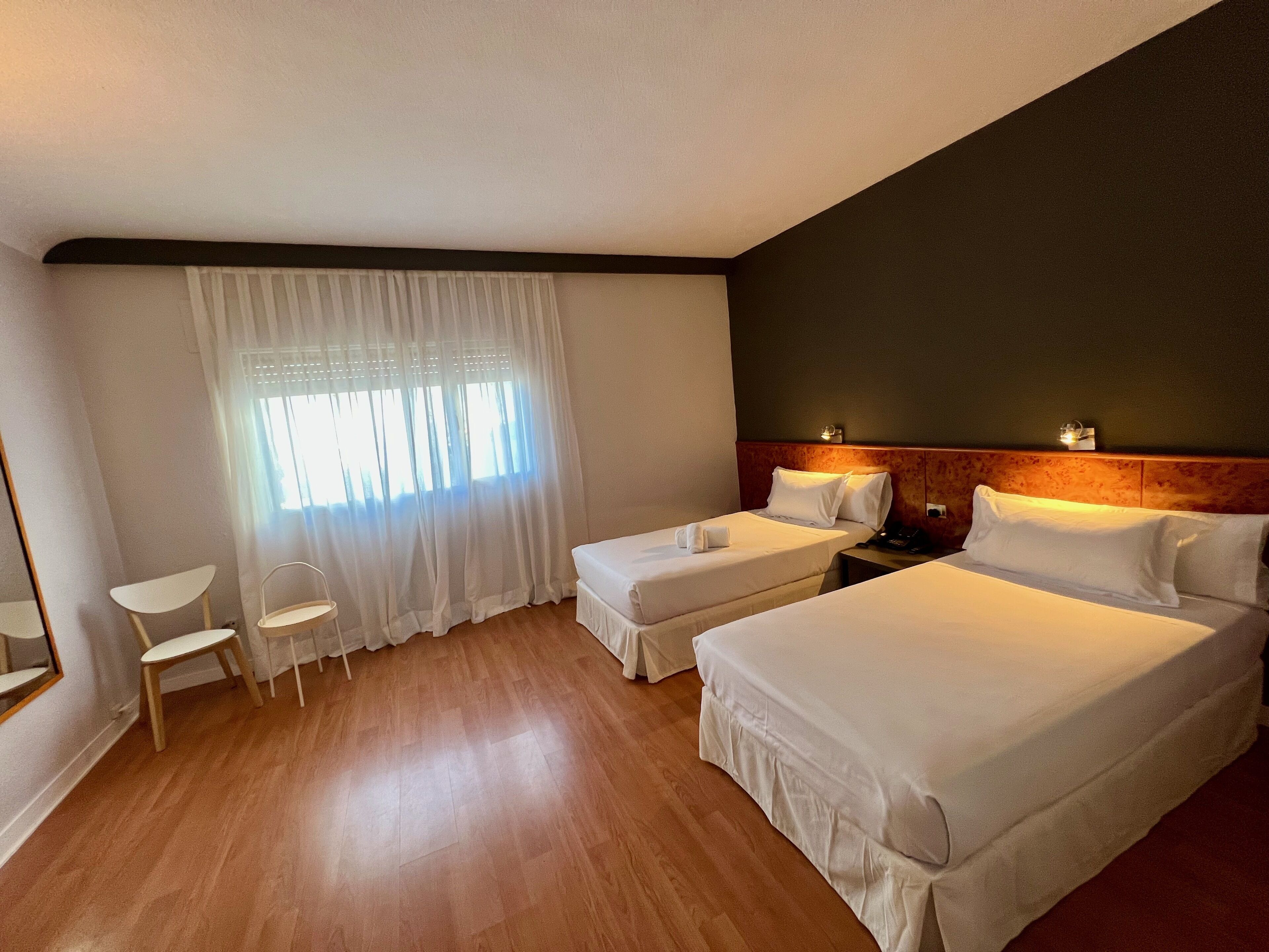 Une chambre d’hôtel avec deux lits simples, une petite table, une chaise et une fenêtre avec des rideaux légers.