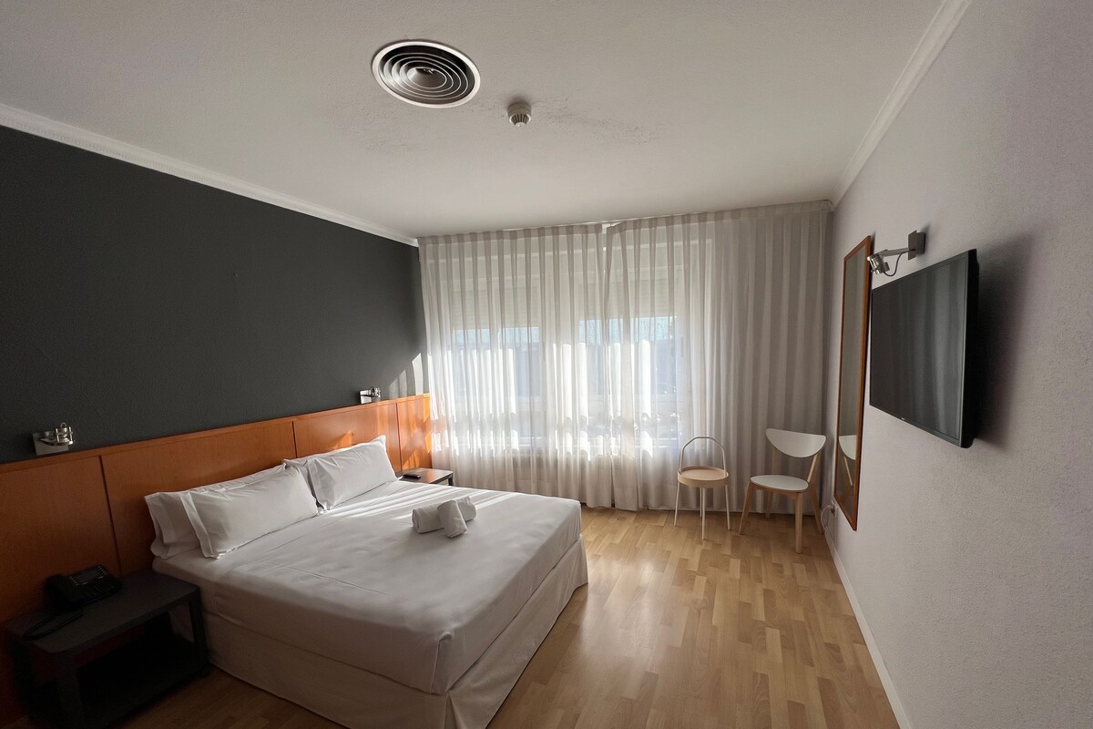 Ein modernes Hotelzimmer mit einem großen Bett, einem Nachttisch, einem Sessel, einem an der Wand befestigten Fernseher und einem Fenster mit transparenten Vorhängen.