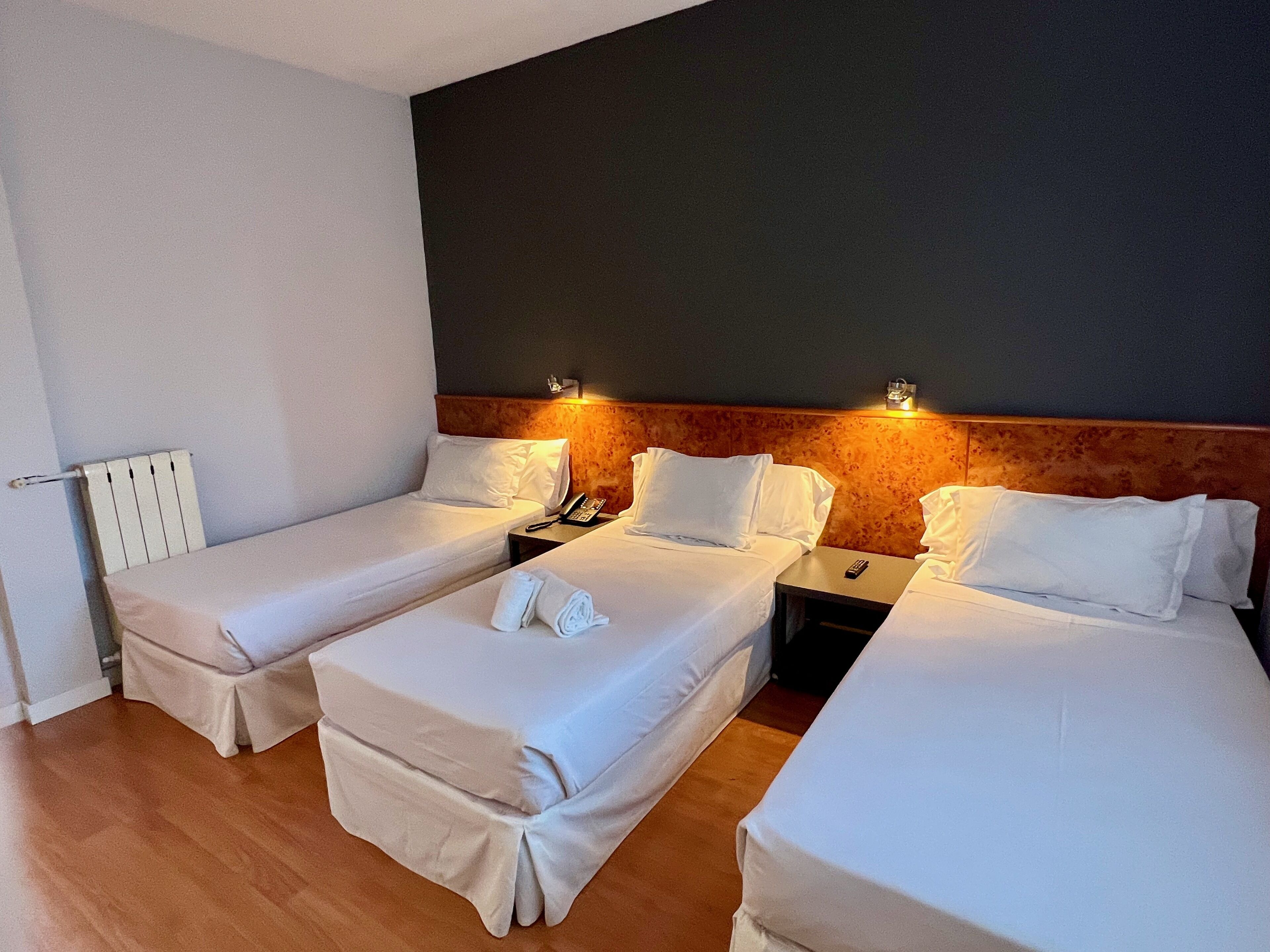 A hotel room with two single beds, a small table, a chair, a bedside table with a lamp, and a wall-mounted phone.