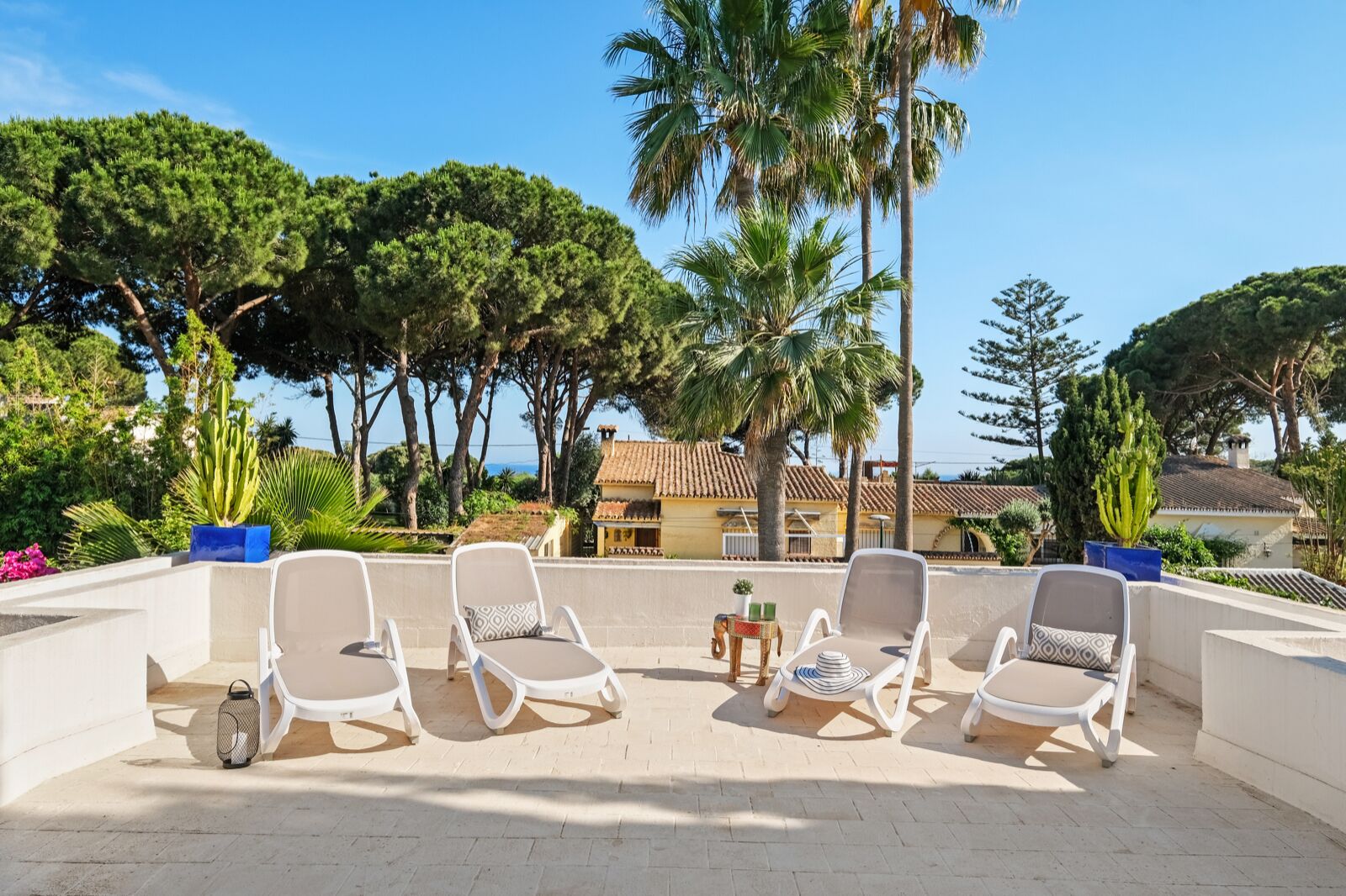 Une terrasse avec quatre chaises longues blanches, une petite table ornée d’une plante, et une vue sur une zone côtière bordée de palmiers et de bâtiments.