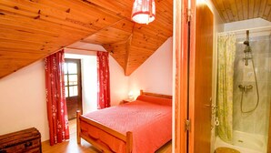 Une chambre avec un plafond en bois, un lit recouvert d’une couverture rouge, une commode en bois et un espace douche doté d’une porte en verre.