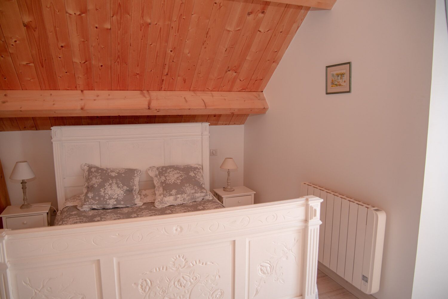 A bedroom with a wooden ceiling, a white headboard, two bedside lamps, and a radiator.