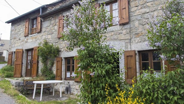 A stone building with wooden shutters, a white table, and flowering plants. - 3/4 pers rental in Grandviala (LA CHAZE-DE-PEYRE)