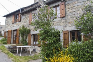 A stone building with wooden shutters, a white table, and flowering plants. - 3/4 pers rental in Grandviala (LA CHAZE-DE-PEYRE)