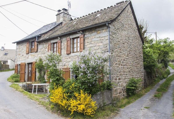 A stone house with a sloped roof, wooden shutters, and a small garden with flowering plants. - 3/4 pers rental in Grandviala (LA CHAZE-DE-PEYRE)