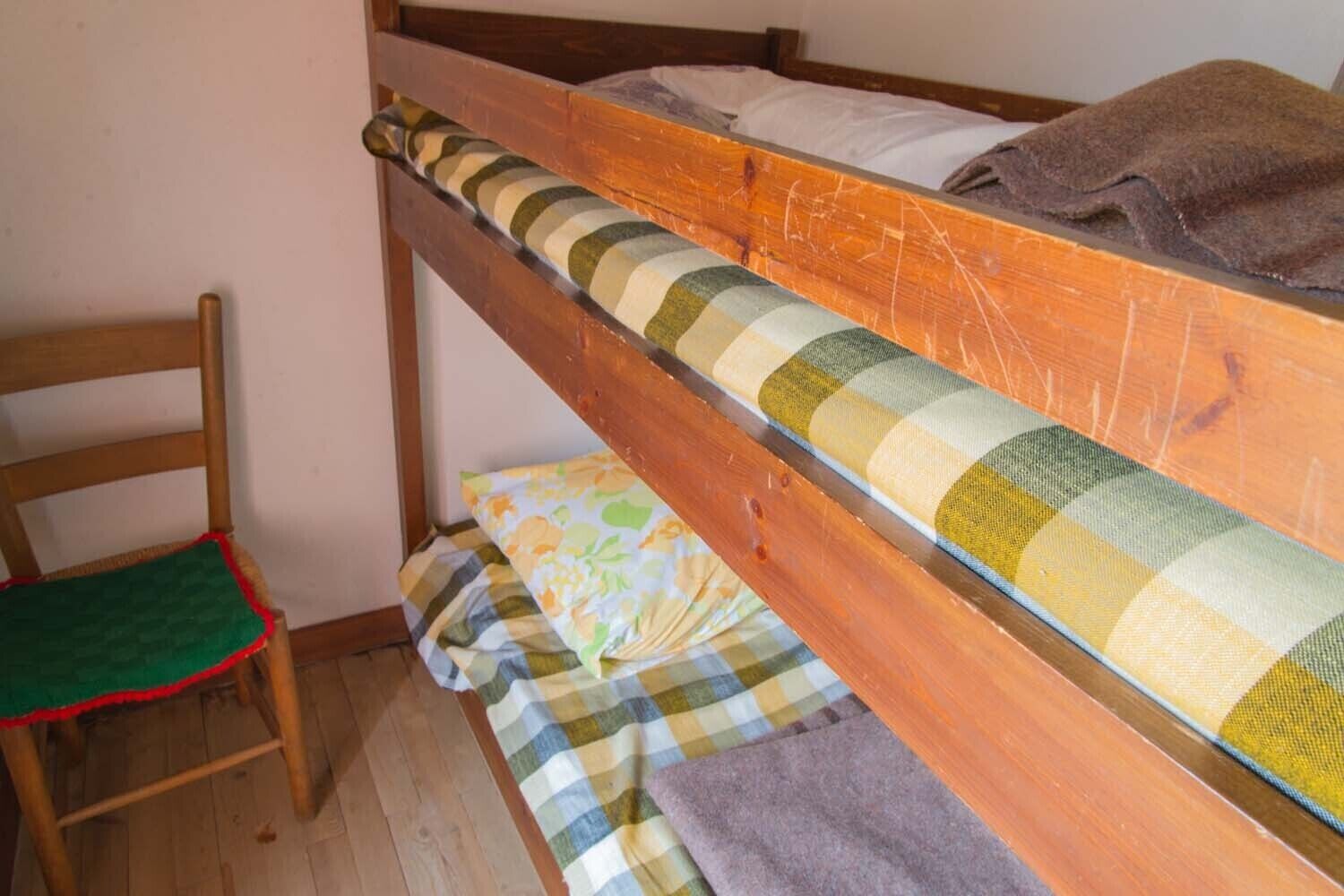 A bunk bed with a wooden frame, a chair with a green seat cushion, and a patterned blanket on the floor.
