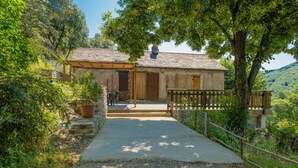 Una casa de piedra rústica con un porche de madera, rodeada de vegetación exuberante y un camino de grava que conduce a la entrada.