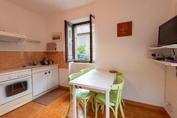 Una pequeña cocina con una mesa blanca y sillas verdes, una ventana que ofrece vistas a un edificio y una estantería con un televisor.