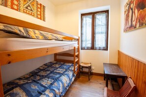 Una habitación pequeña y acogedora con dos literas, una mesa de madera, un taburete pequeño y una ventana con cortinas estampadas.