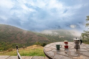 A stone table with a cup, a thermos, and a kettle, overlooking a mountainous landscape with a village below.