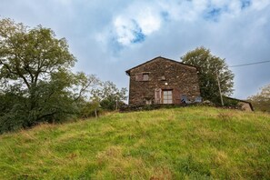 Une maison en pierre avec un toit en pente, une porte et une fenêtre, entourée d’herbe et d’arbres.