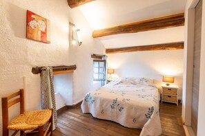 Une chambre avec un plafond en bois, un lit recouvert d’une couvre-lit à motifs floraux, une chaise, une table de chevet avec une lampe et une fenêtre.