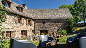 Un bâtiment en pierre doté d’un toit en pente, de grandes fenêtres et d’une entrée en pierre. Il y a des espaces extérieurs aménagés avec des chaises et des tables.