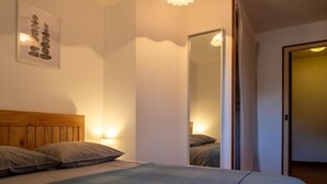 Un dormitorio con cabecera de madera, cama con ropa de cama gris, luz fijada en la pared y un espejo que refleja la habitación.