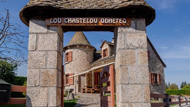 Un arc en pierre portant l’inscription « LOU CHASTELOU ODHETO » mène à un bâtiment traditionnel en pierre, surmonté d’un toit conique.