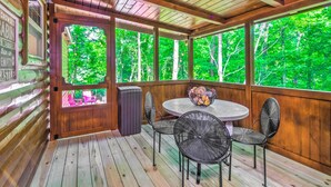 Una terraza de madera con una mesa redonda y cuatro sillas, rodeada de paredes de madera y amplias ventanas que dan a un bosque.