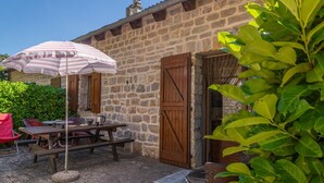 A stone building with a wooden door, a picnic table with chairs, and a striped umbrella. - Gîte "Le Noyer" 4 pers in Blajoux (BLAJOUX)