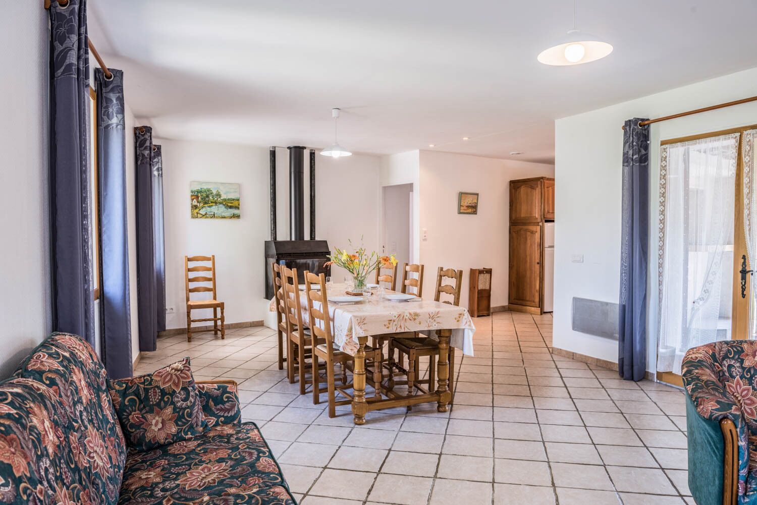 Ein Essbereich mit einem Tisch für vier Personen, umgeben von Holzstühlen. Im Vordergrund befindet sich eine Couch, und im Hintergrund ist eine Küchenzeile zu sehen.