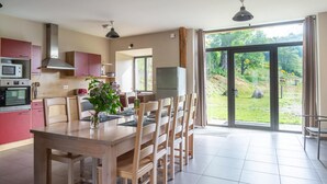 A modern kitchen with red cabinets, a wooden dining table, and a view of a garden through large sliding doors. - Lzg090a - Gîtes de France 3 Épis 6 Pers à Coulagnettes (SAINT-AMANS)