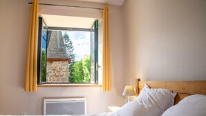 A bedroom with a bed, a nightstand, a lamp, and a window with curtains. - Lzg090a - Gîtes de France 3 Épis 6 Pers à Coulagnettes (SAINT-AMANS)
