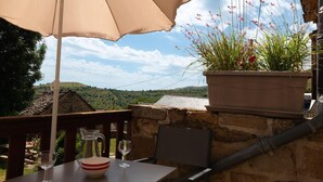 A table with a beige umbrella, a wine glass, and a bowl, set against a stone wall with a potted plant and a view of hills.