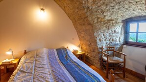 A bedroom with a stone ceiling, a bed with a striped blanket, a wooden chair, and a nightstand with a lamp.