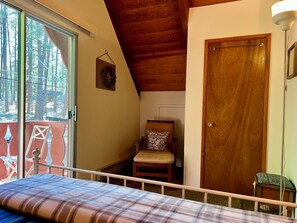 A bedroom with a bed, a wooden door, a chair, and a window overlooking a forest. - Creekside Chalet - Prescott Cabin Rentals (Prescott)