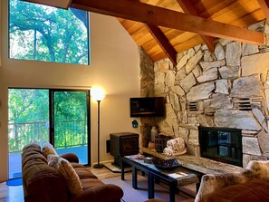 A living room with a stone wall, a fireplace, a TV mounted on the wall, and a large window overlooking a deck. - Creekside Chalet - Prescott Cabin Rentals (Prescott)