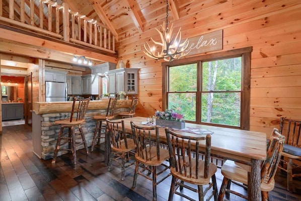 A rustic cabin interior with a stone fireplace, wooden dining table, and a large window offering a view of the forest.