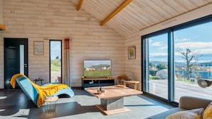 Ein modernes Wohnzimmer mit Holzbalkendecke, einem großen Fernseher, einer Couch, einem Couchtisch und Blick auf eine Landschaft durch große Fenster.