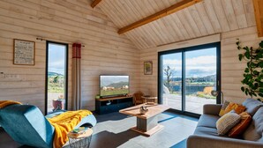 Ein gemütliches Wohnzimmer mit Holzbalkendecke, einem Fernseher, einem Sofa, einem Couchtisch und Blick auf die Landschaft durch große Fenster.
