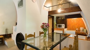 A dining area with a glass table, wicker chairs, a vase with flowers, a clock, and a kitchen with orange tiles and appliances. - House (SAINTE-ENIMIE)