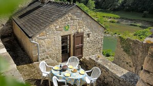 A stone building with a tiled roof, a wooden door, and a table with chairs outside. - House (SAINTE-ENIMIE)