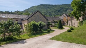 Ein ländliches Steinhaus mit befestigter Auffahrt, umgeben von Grünflächen und einem Hügel im Hintergrund.