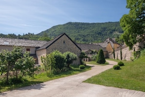 A rural stone building with a paved driveway, surrounded by greenery and a hill in the background.