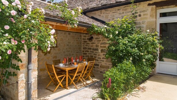 Ein Gebäude aus Stein mit einem Ziegeldach, einem Holztisch mit Stühlen und einem Garten mit blühenden Pflanzen.