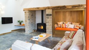 Ein gemütliches Wohnzimmer mit Kamin aus Stein, einer Couch mit bunten Kissen, einem Flachbildfernseher und einem kleinen Tisch mit Orangen und einer Zeitschrift.