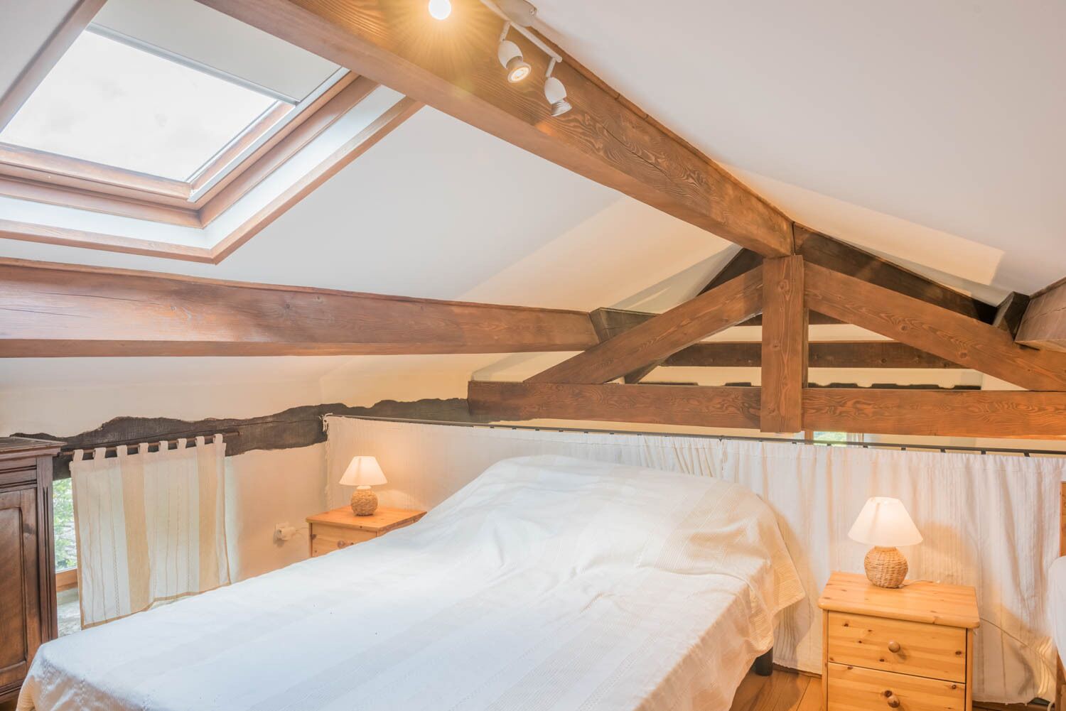 Una camera da letto con lucernario, travi in legno, un letto, due comodini con lampade e una finestra con tende.