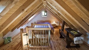 A high-ceilinged attic bedroom with a sloped roof, wooden beams, a bed, a nightstand, a chair, and a small table.