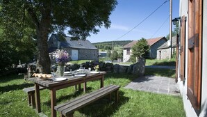 A stone building with a stone pathway leading to a wooden table set for a meal, surrounded by grass and trees.