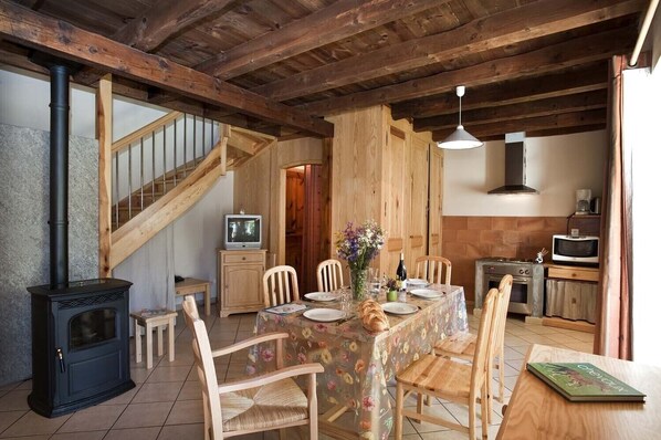 A rustic dining area with a wooden table, chairs, and a television. A wood-burning stove is visible on the left, and a kitchen area with a microwave and stove is on the right. - Lzg129 - Gîtes de France 3 Épis 6 Pers. à Saint Paul le Froid (SAINT-PAUL-LE-FROID)