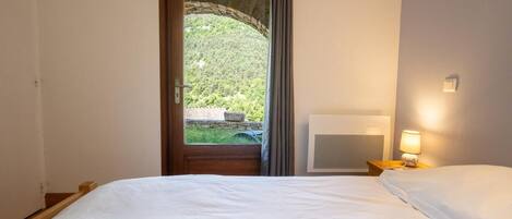 Quarto com cama, mesa de cabeceira com abajur, aquecedor na parede e vista para uma paisagem verde pela janela.