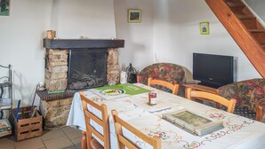 A dining area with a stone fireplace, wooden chairs, a table with a tablecloth, a television, and a staircase.