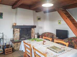A dining area with a stone fireplace, wooden chairs, a table with a tablecloth, a television, and a staircase.